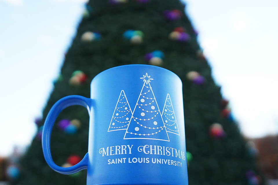 A coffee mug that says "Merry Christmas Saint Louis University" with an image of three trees sits in front of a large Christmas Tree on campus.