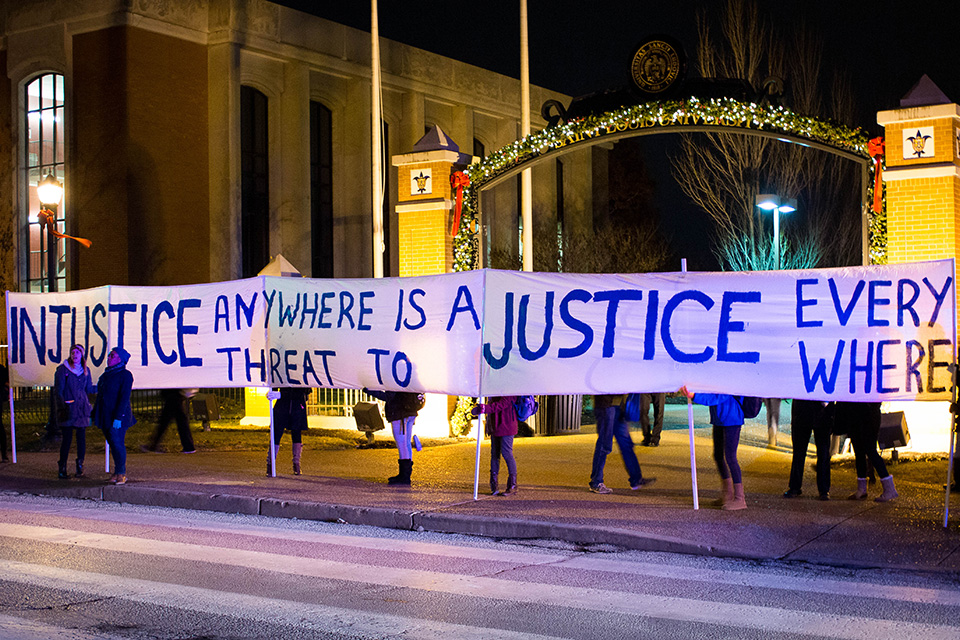 Students stand along a street on campus in front of an archway decorated by Christmas lights. They hold a long banner that reads "Injustice Anywhere is a Threat to Justice Everywhere"