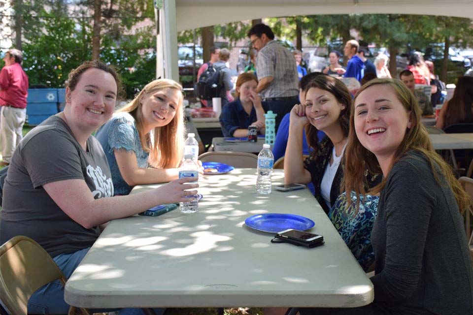 Four students sit at an outdoor table, with bottled water and paper plates in front of them.