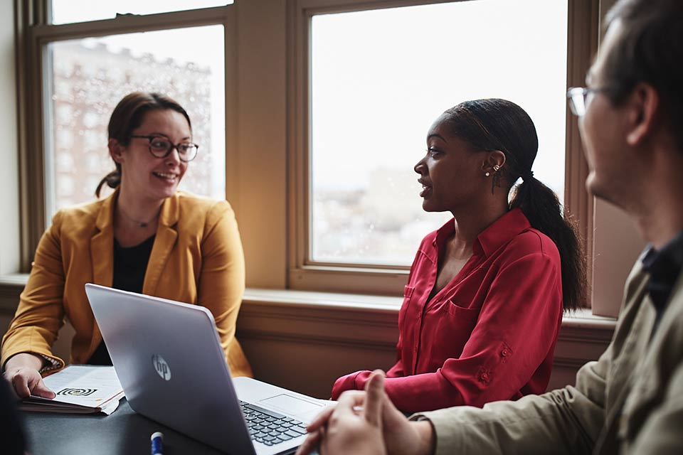 Three students sitting around a table with a laptop, and having a discussion.