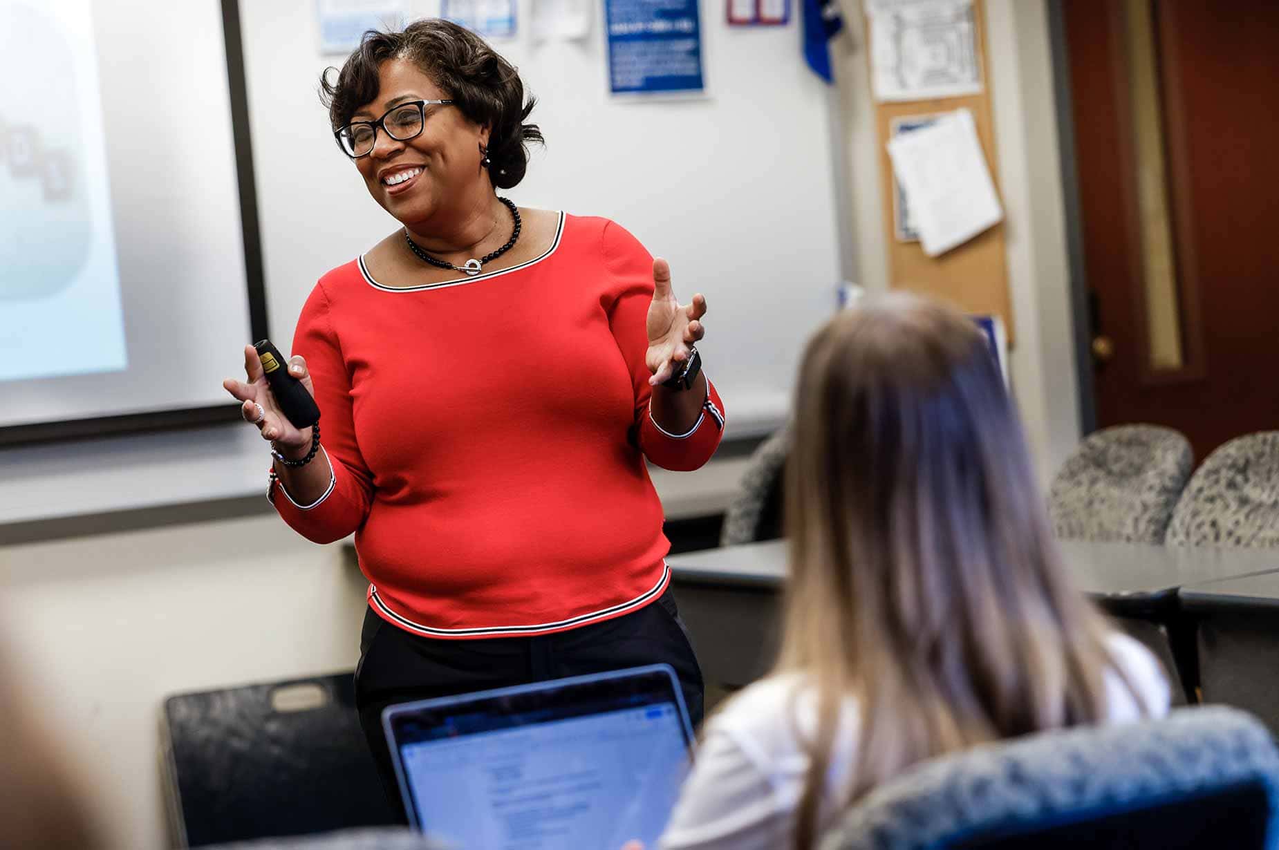 A female faculty member stands in front of a classroom of students. She smiles to the classroom.