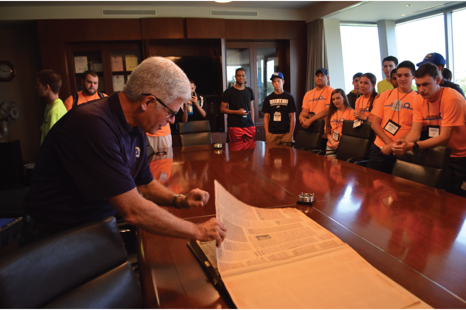 Students in orange standing around a table in a conference room as part of Allsup Entrepeneurship Academy