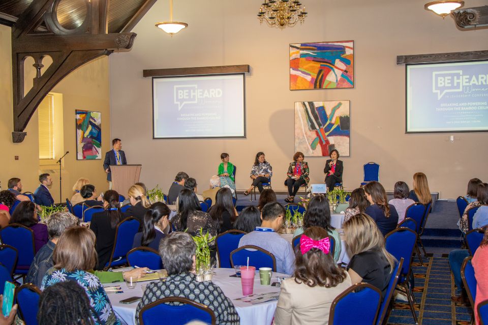 A crowded room of conference attendees are seen from behind while sitting at tables, watching while four speakers sit on a stage and one stands at a lectern.