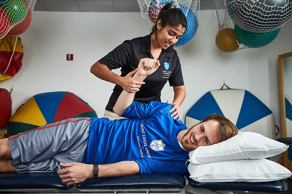 A physical therapy student assists a patient lying on a table with an arm exercise, with physical therapy equipment in the background.