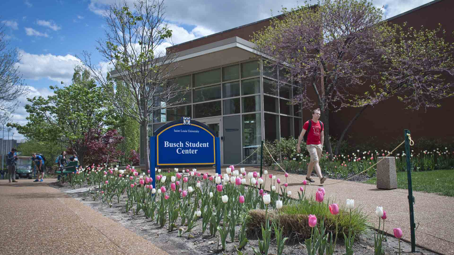 Exterior of the Busch Student Center with a student walking by and flowers blooming.