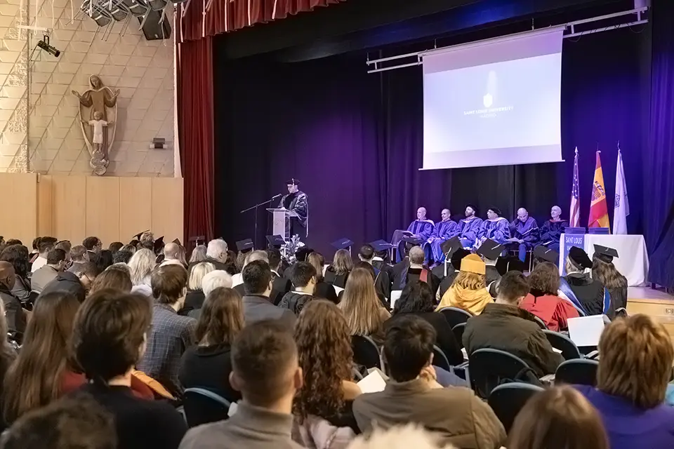 Man speaks on stage in front of podium to an auditorium full of graduating students and other attendees present.