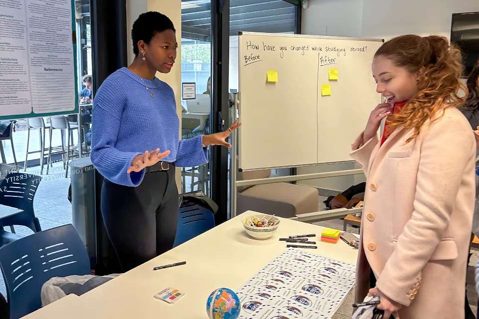 A girl explains a psychology project to a fellow student through interactive activities at a stand with a large poster behind her. 