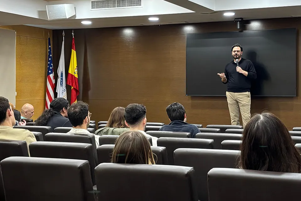 People sitting in brown auditorium chairs look at the speaker on stage who is speaking into a handheld microphone with the American and Spanish flags on the stage to the left.