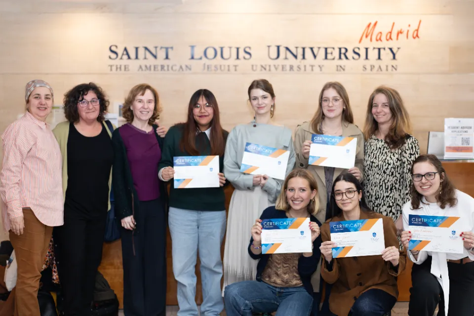 Three women standing look onward at camera next to three students posing with certificates standing in the same row. There are three other students posing with the same certificate who are kneeling with a woman standing behind them. There is a logo on the wall in the background.