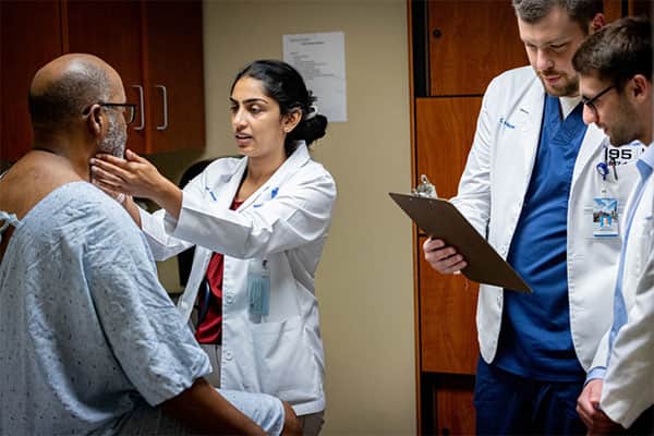 A resident examines a patient in a hospital gown in an examination room while two other residents review his chart.