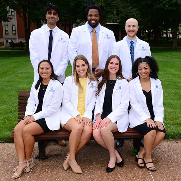 A group of medical students wearing their white coats sit on a bench outside of College Church. They are posed and smiling at the camera.