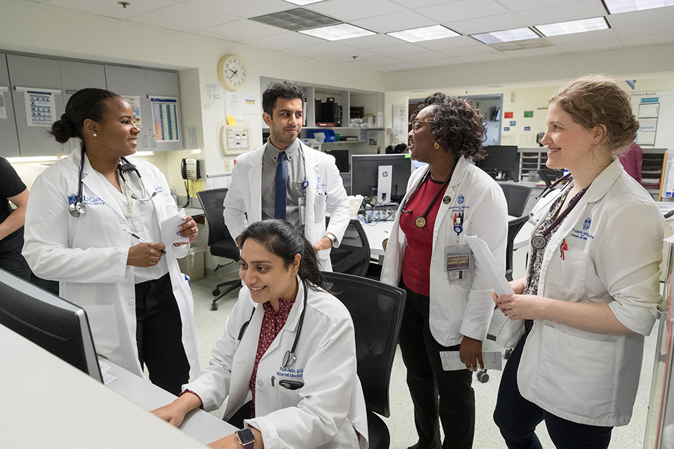 Four medical students stand behind a classmate who is seated and working at a computer. Students are smiling.