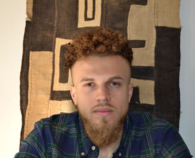 Art historian Bentley Brown gazes directly at the viewer while sitting in front of a brown and beige swath of fabric with an abstract design.