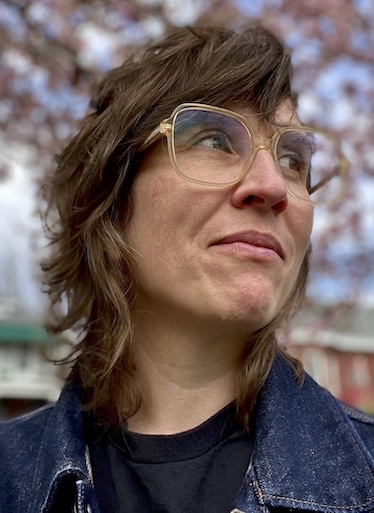 Jessica Baran Curator Jessica Baran, wearing a blue jean jacket and large-framed eyeglasses, grins as she looks up and to the right