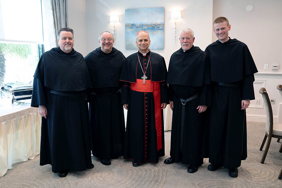 A photo of five men dressed in black robes taken inside a room. Pope Leo XIV, middle, wears a black and red rob with a red zucchetto on his head. He also wears a cross and a white clerical collar. 