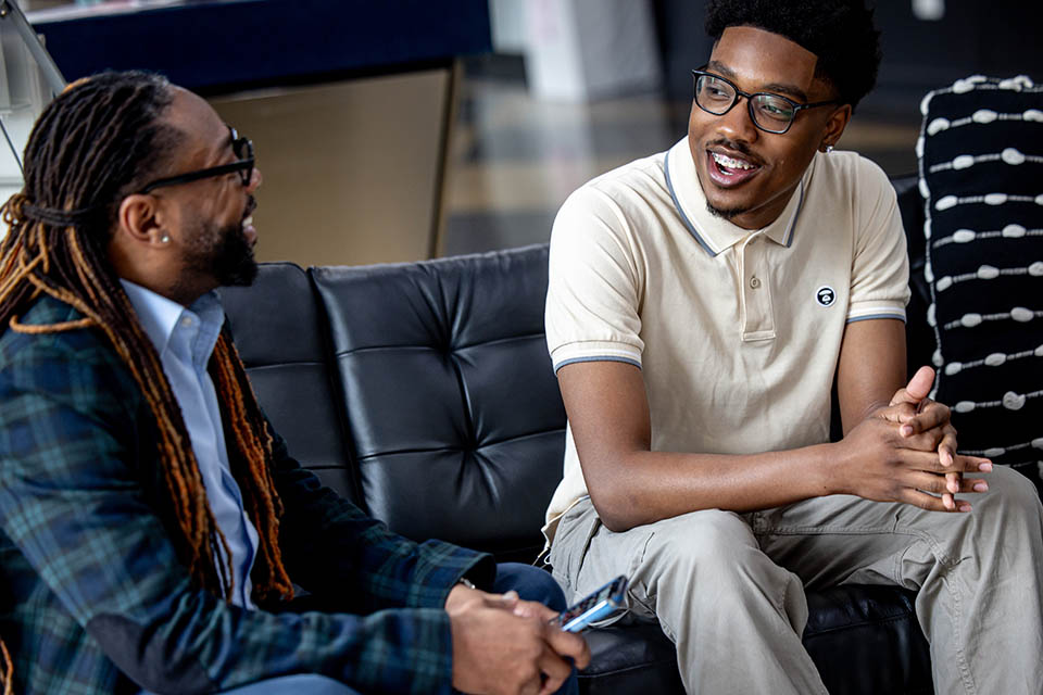 Dr. Richard Marks, left, meets with student Russell Hooker Jr. The pair are sitting down on a black couch inside a room engaged in a conversation.