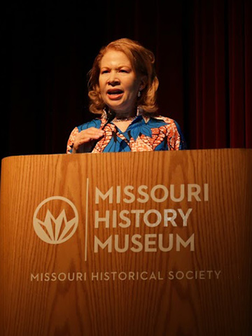 Angie Clay Thomas wears a blue and orange floral outfit as she speaks behind a podium labeled Missouri History Museum against a deep red curtain.
