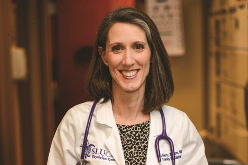 Dr. Kimberly Schiel poses for a photo in a dimly lit room in a white coat and a stethoscope around her neck.