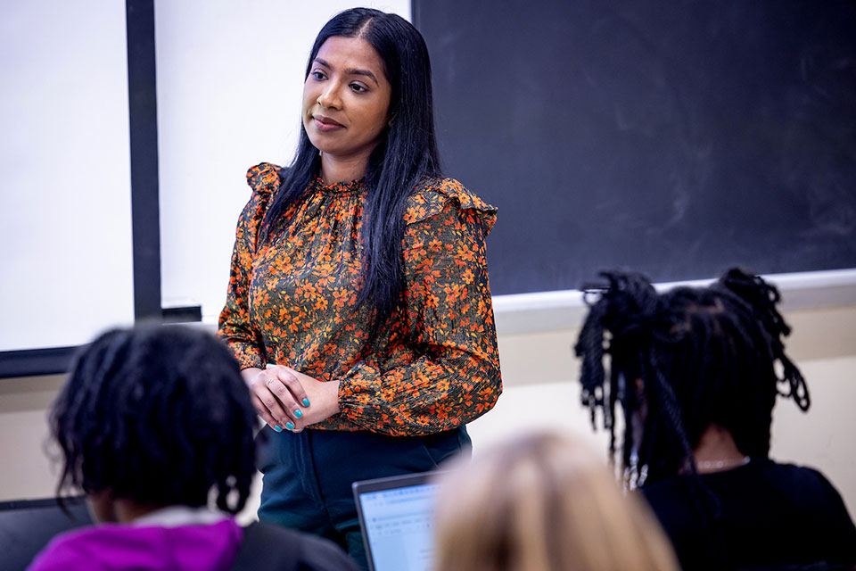 Professor Popy Begum, Ph.D., wearing a floral blouse, leads a classroom discussion. Students, some of whom have laptops, focus on her.