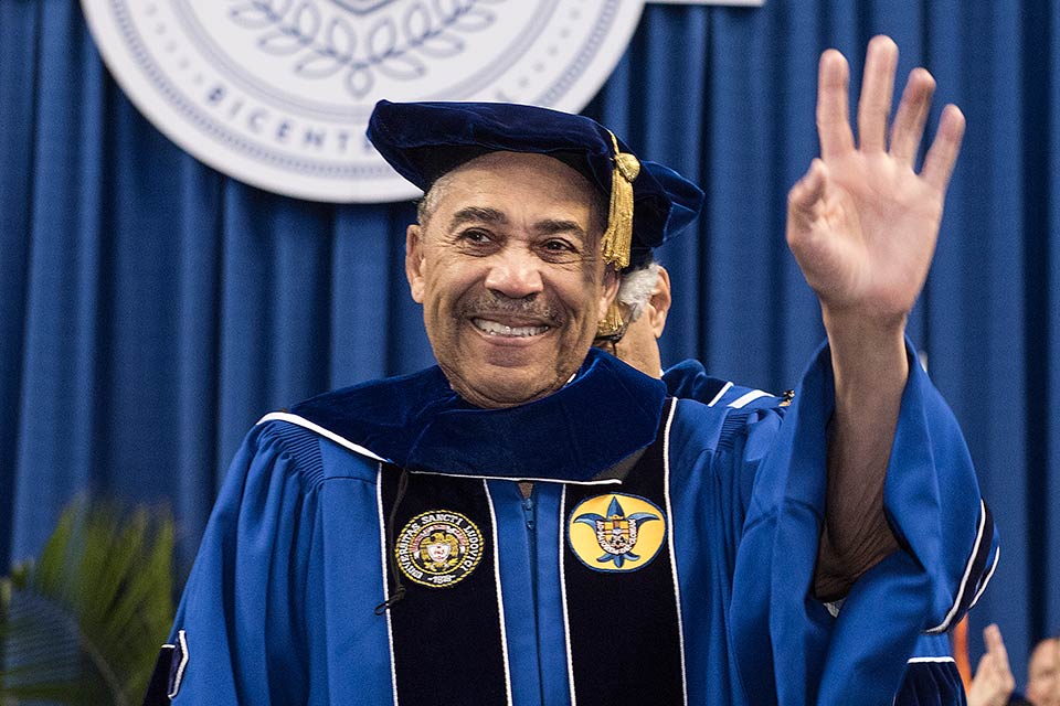 William L. Clay, dressed in a blue academic gown, waves enthusiastically at the crowd during the 2018 commencement ceremony at Saint Louis University. 