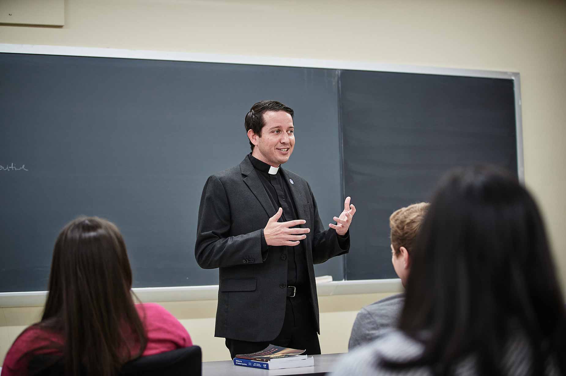 A Jesuit faculty member speaks to students in front of a classroom with a blackboard. The heads of students can be seen in the foreground.