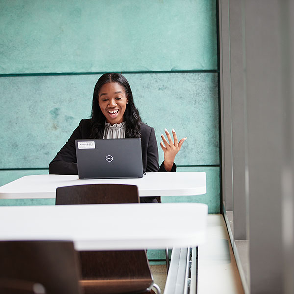 A student sits at a table and video conferences from her laptop. She’s smiling and speaking to the screen.