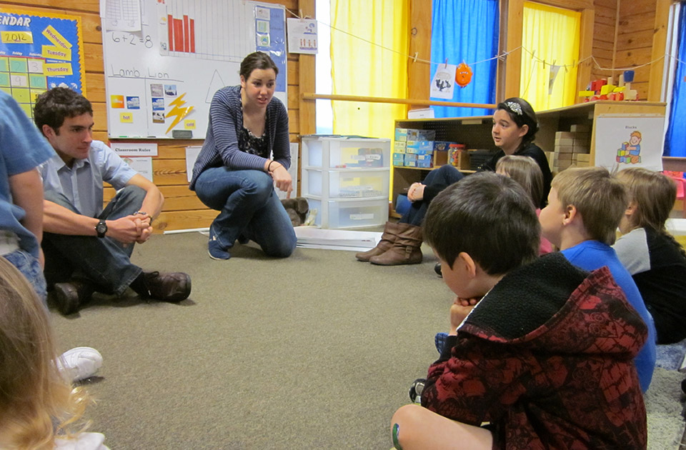 Students work at local school. Two SLU students sit on the floor at the front of a classroom with younger students gathered in a semi-circle.